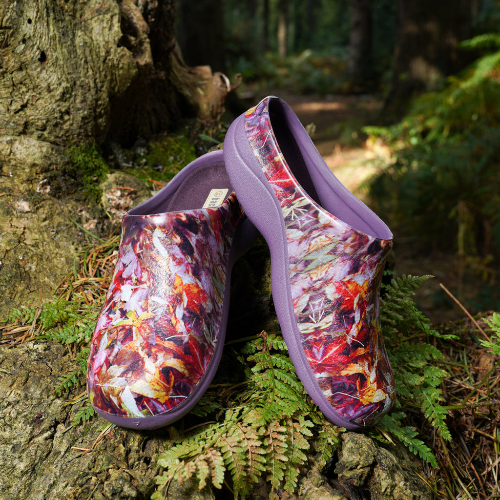 Pair of purple forest leaf-patterned gardeningshoes on a forest floor with trees and ferns in the background.