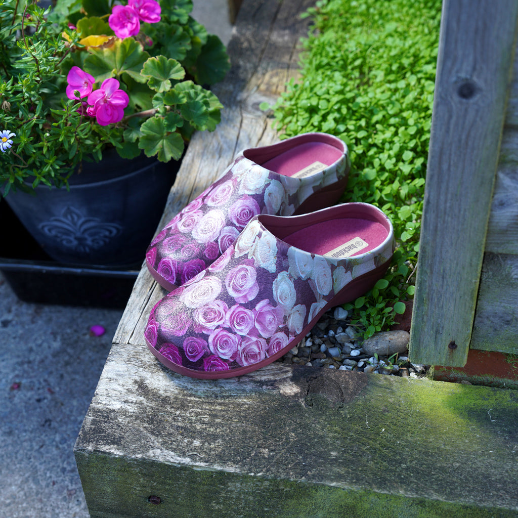 Purple floral-patterned clogs on a wooden planter in a gardening setting with pink flowers in the background